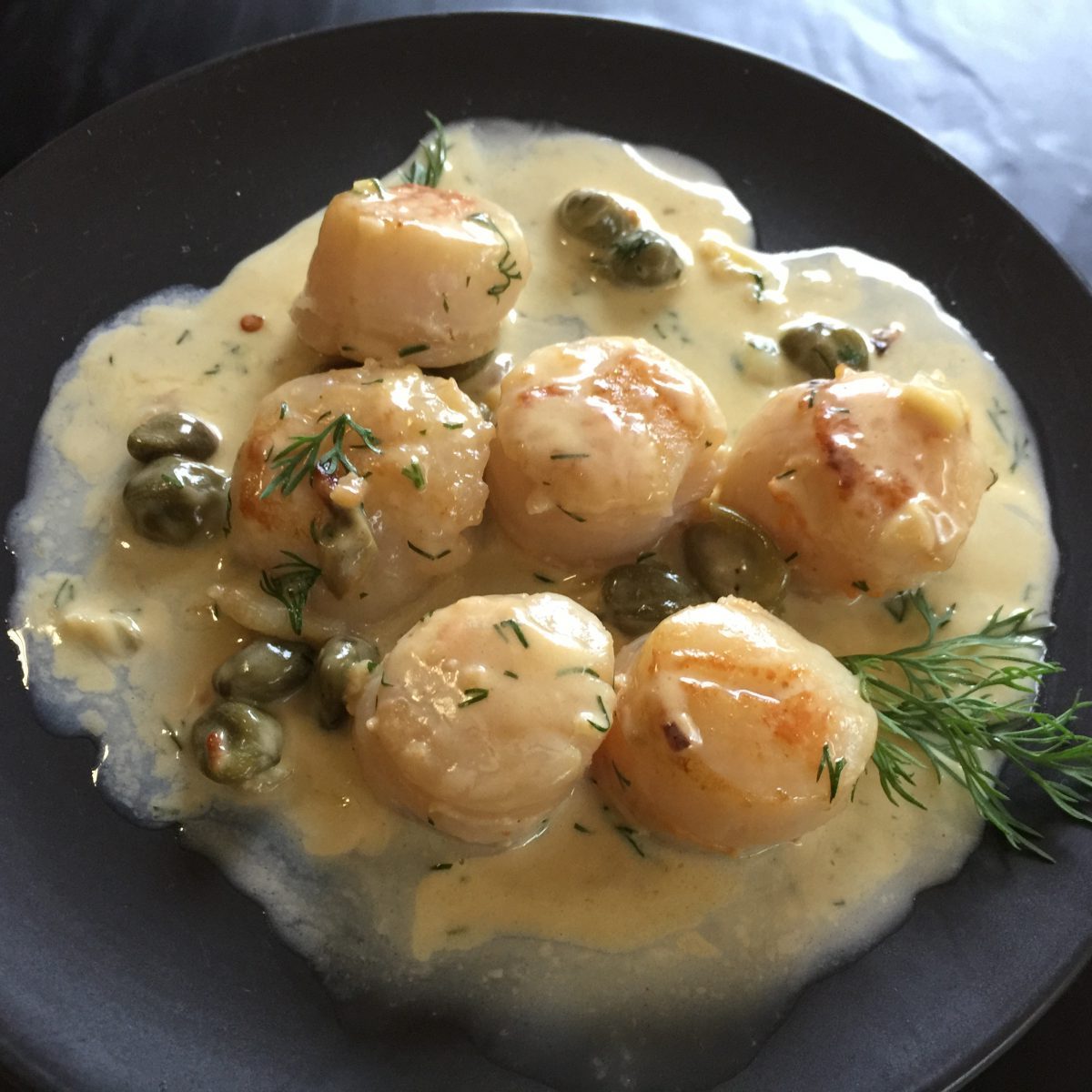 6 Scallops with Lemon Dill and Caper Sauce served on a black plate by cookingmealsforone.com
