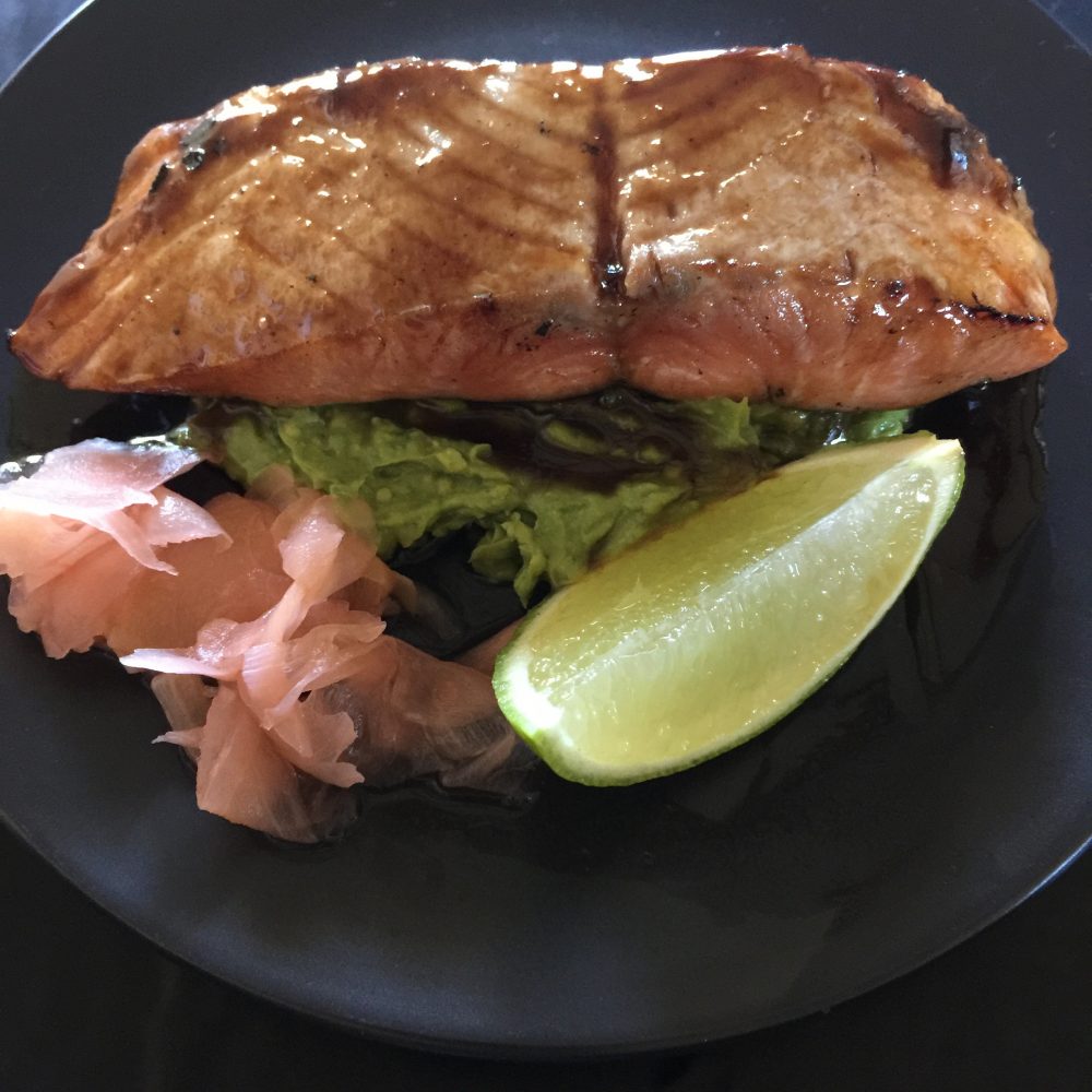 A fillet of smoked salmon dressed in a delicious teriyaki sauce served with smashed wasabi avocado, pickled ginger with a wedge of lime on a black plate by cooking meals for one
