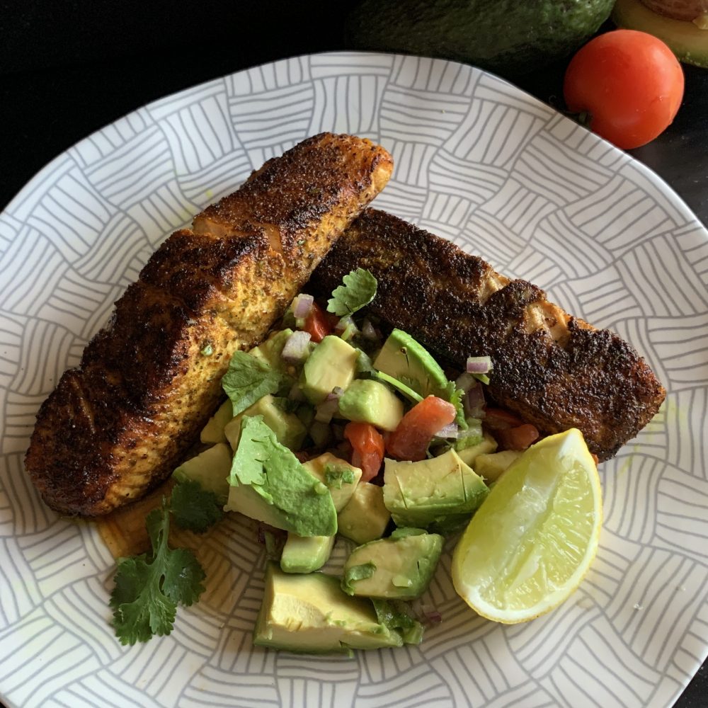 2 juicy salmon fillets coated in Moroccan Spices and pan seared in melted butter until blackened and just cooked. Served with an Avocado Salsa of avocado chunks chopped red onion cherry tomatoes coriander and dressed in lime juice Served on a patterned white plate with a wedge of lime
