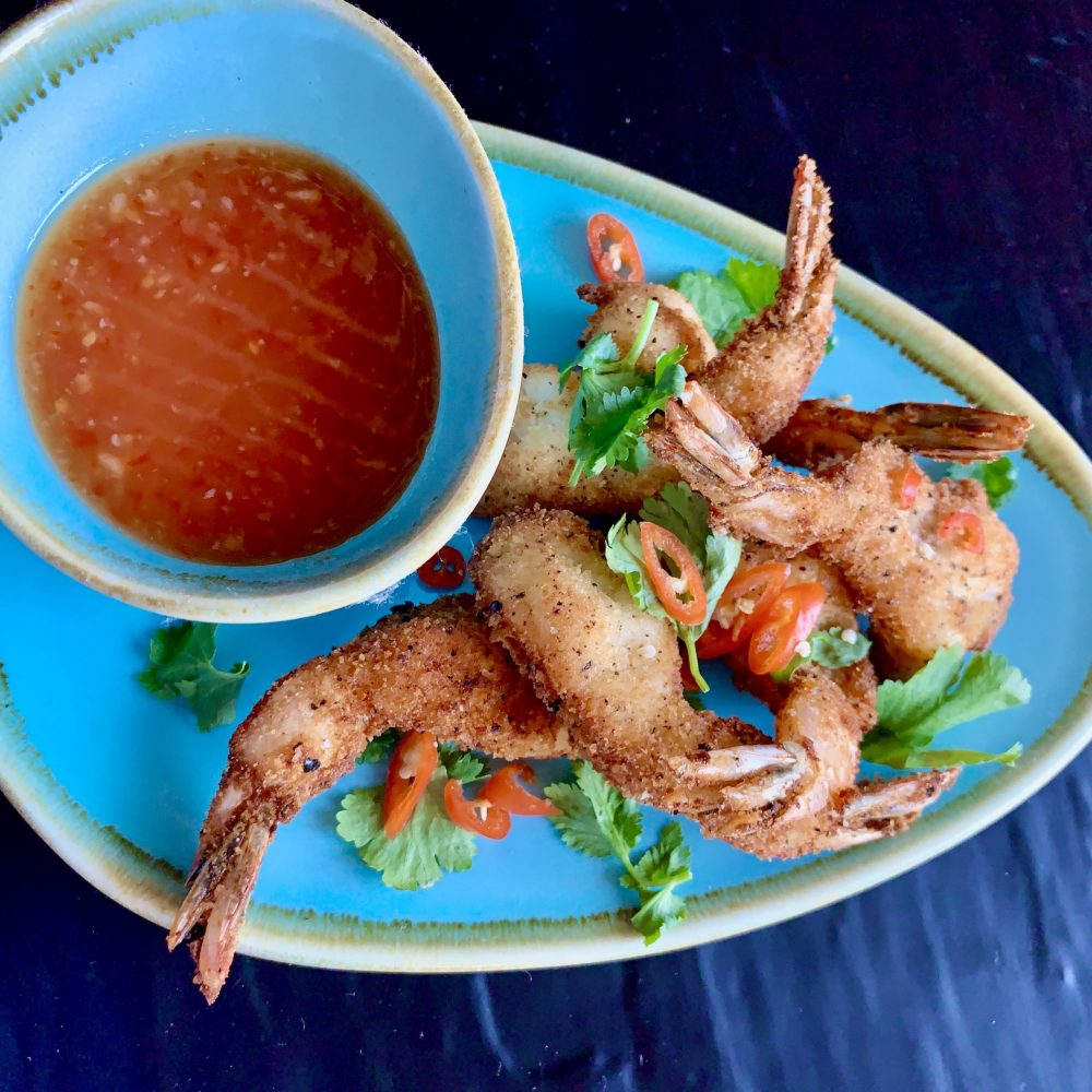 These crispy crunchy piri piri spiced salt and pepper crumbed prawns cutlets are garnished with coriander leaves and slices of red chilli and served on a blue oval plate with a blue smaller bowl of my "Thai sweet chilli sauce for dipping