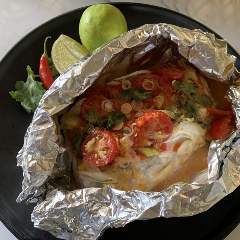 A teamed fish parcel of ssucculent flavoured fish, topped with sweet chilli sauce, fish sauce, lime juice, sliced green shallots, grated ginger and halved cherry tomatoes with a sprig of coriander leaves, wrapped in foil and cooked ion oven. Served on a black plate with a wedge of lime