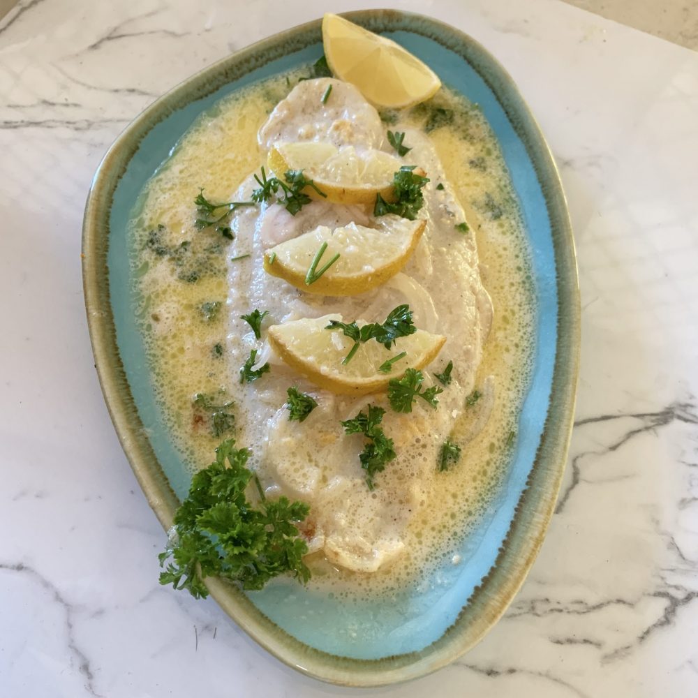 Creamy Lemon Baked Fish For One: A white fillet of fish cooked in a buttery, garlic, mustard flavoured coconut creamy sauce,topeed with freshly chopped parsley. Served on a blue plate with a wedge of lemon. by cookingmealsforone.com