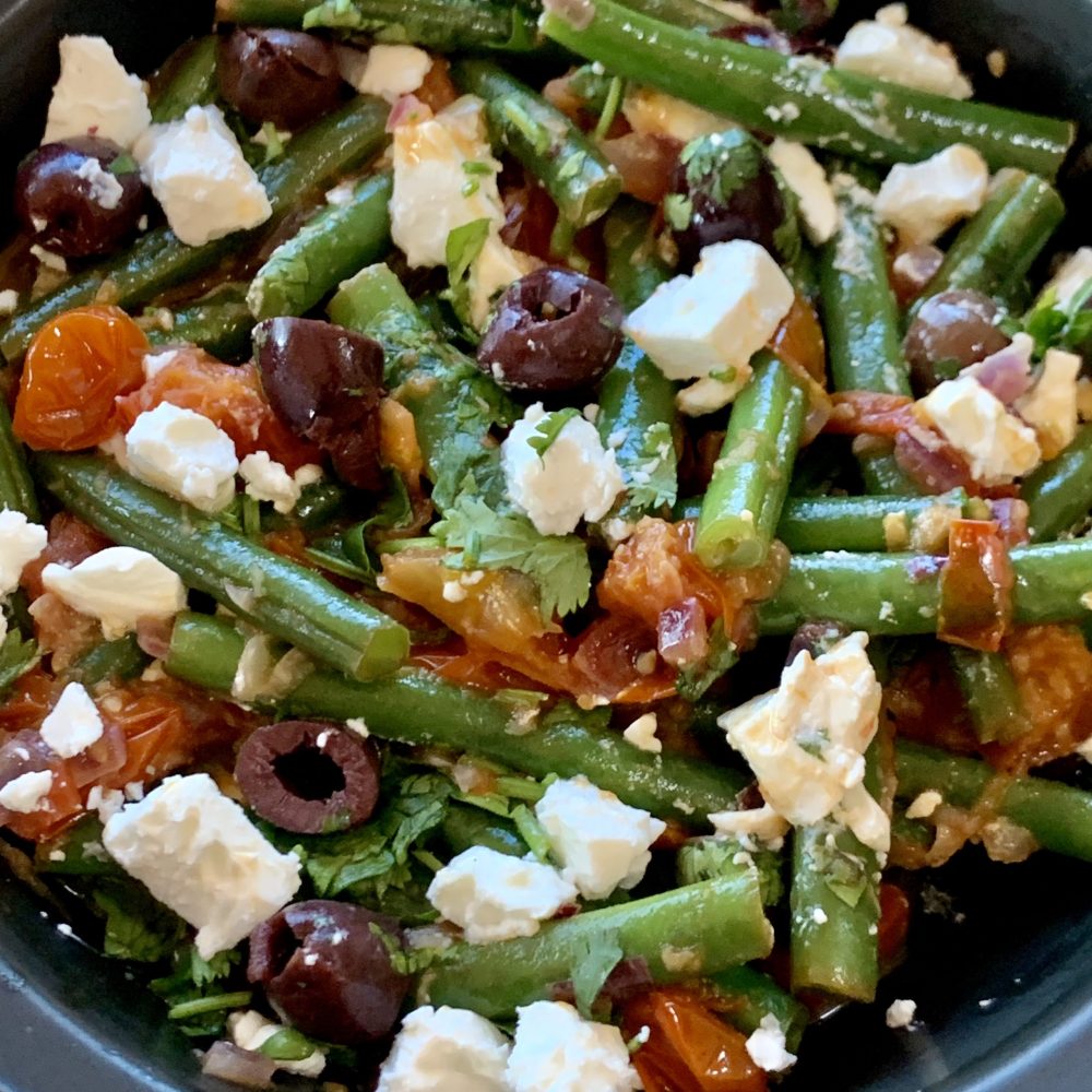 A delicious combination of cooked green beans in a garlic, onion and tomato sauce and garnished with chopped kalamata olives, parsley and crumbled feta served in a black bowl. by cookingmealsforone.com